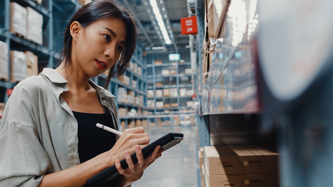 A woman writes on a tablet while looking at items on a shelf in a warehouse